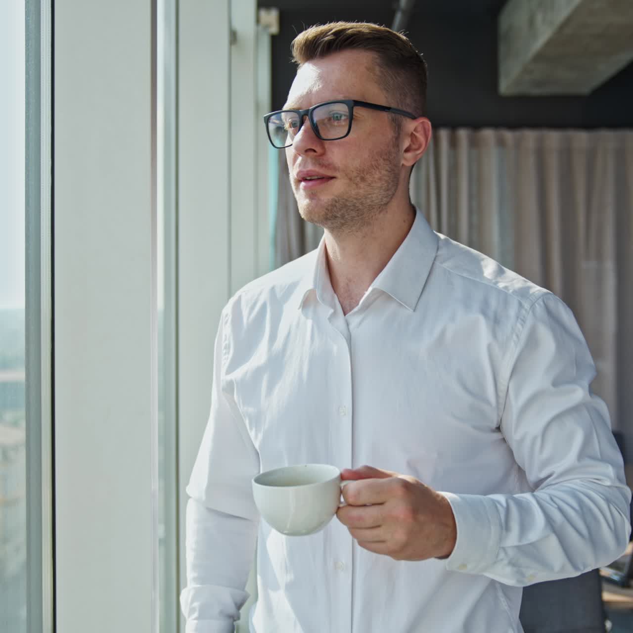 Businessman drinking coffee standing in the spacious office. Man enjoys coffee break looking at the city from a big panoramic window