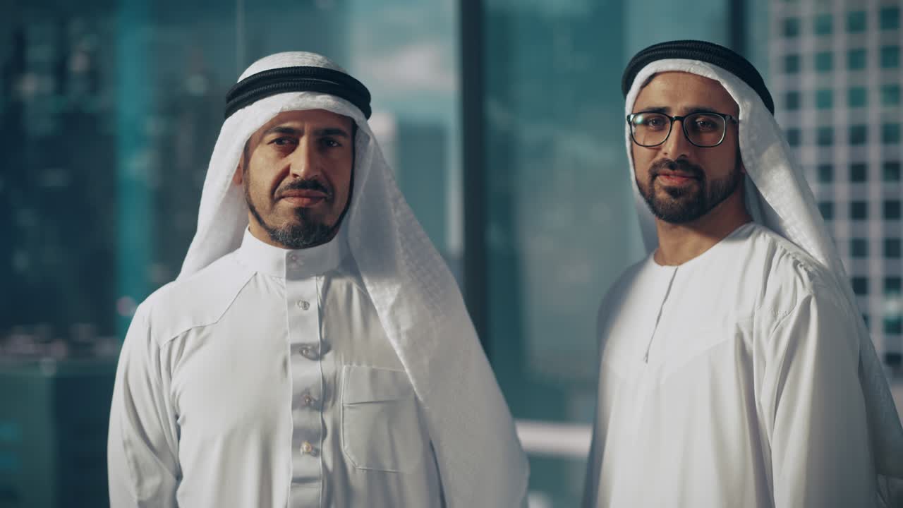 Ritratto di due uomini d'affari musulmani di successo in abiti tradizionali che sorridono dolcemente, indossando una kandura bianca e un agal nero che tiene ferma una ghutra. Concetto di uomo d'affari saudita, emiratino, arabo.