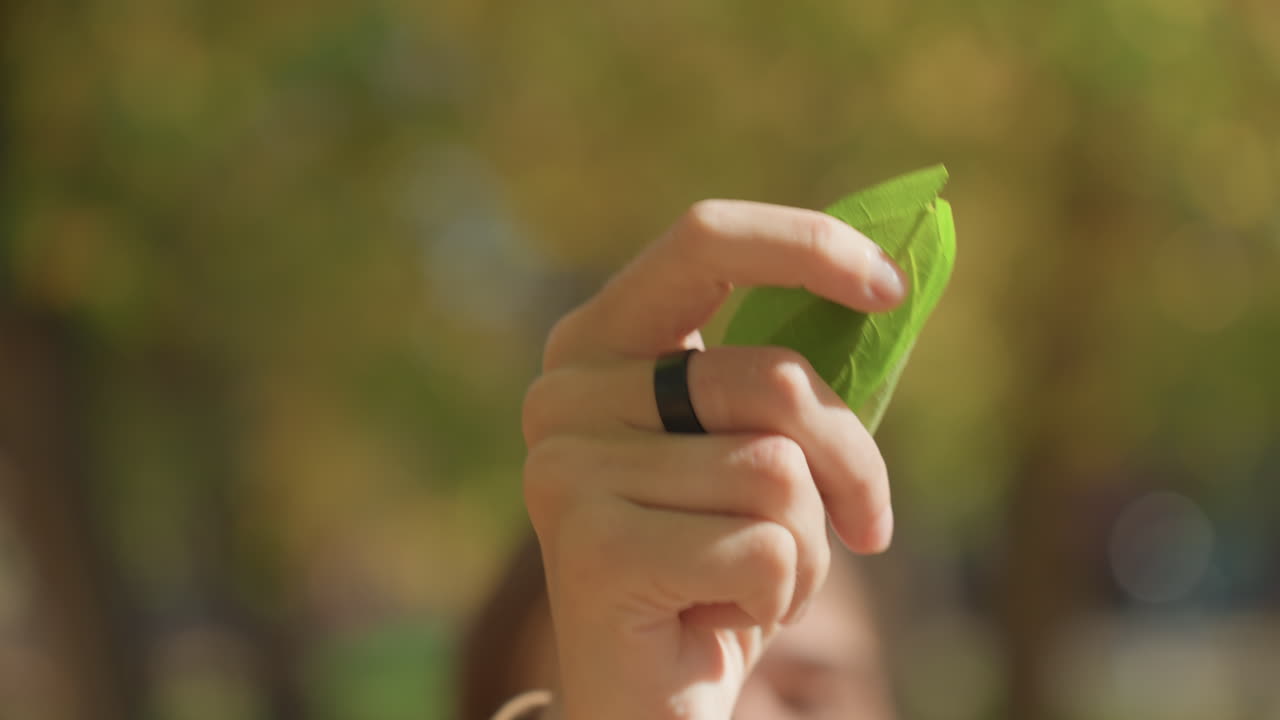 Hand touching green leaf near baby, family nature discovery with mother demonstrating texture while infant watches, closeup of fingers, bracelet and beanie, warm bokeh background and soft autumn light