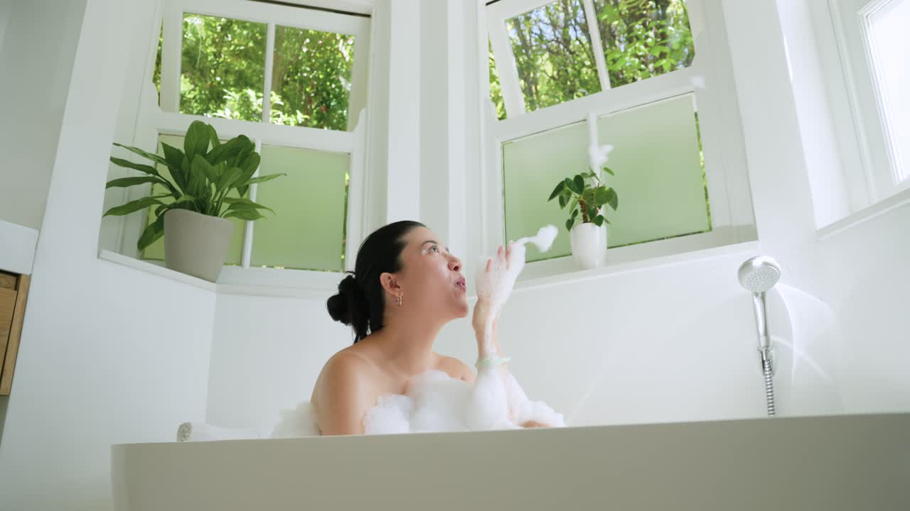 African American woman relaxing in freestanding tub under bay windows, scooping and blowing foam