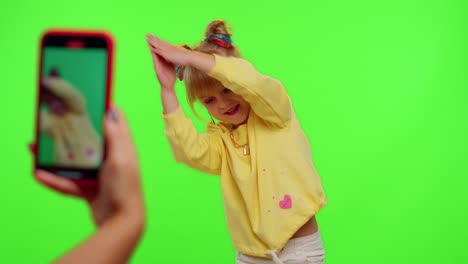 Cheerful-funny-little-teen-girl-kid-dancing-at-camera-filming-video-using-phone-on-chroma-key-wall