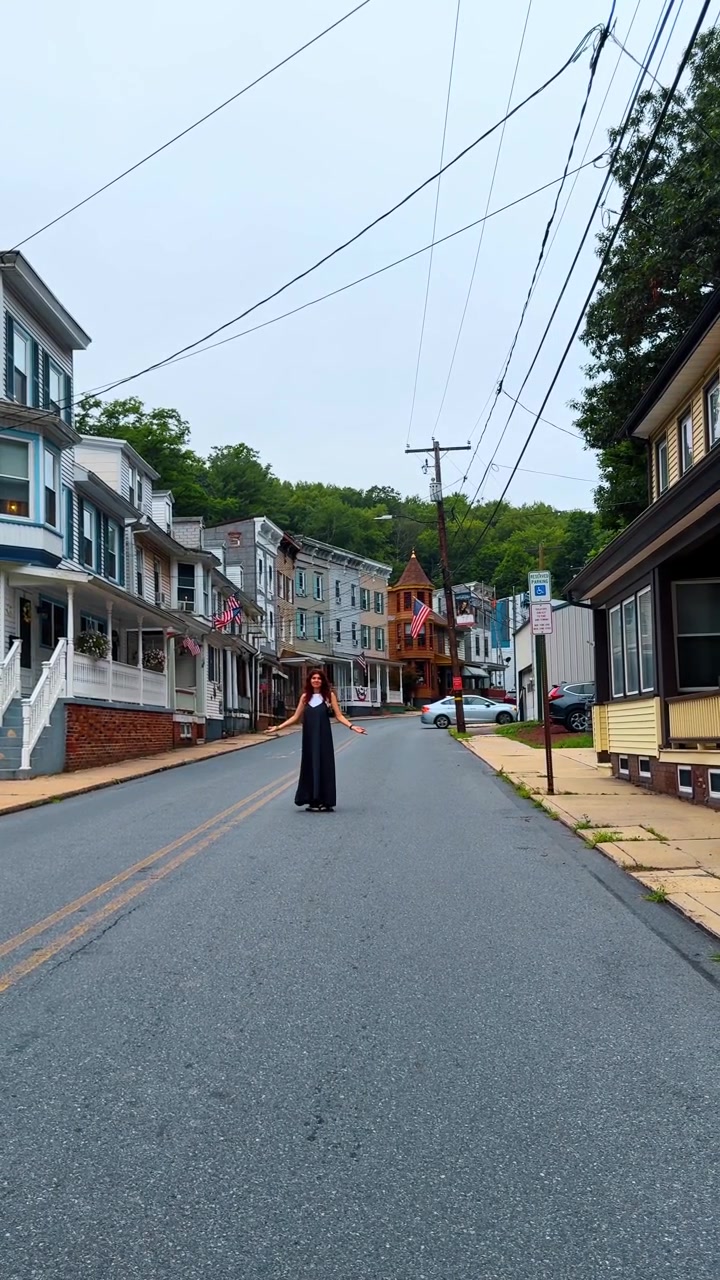 Kvinde i sort kjole går på gaden i Jim Thorpe, Pennsylvania. Kvinde går ned ad en brolagt vej flankeret af farverige historiske huse og amerikanske flag under en overskyet himmel.