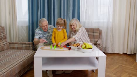 Senior-grandparents-with-child-kid-granddaughter-playing-game,-riding-toy-train-on-railway-at-home