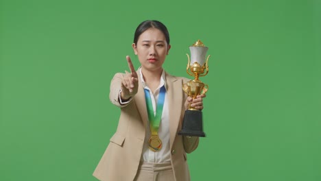 asian business woman in a suit with a gold medal and trophy disapproving with no index finger sign and smiling to camera on green screen background in the studio