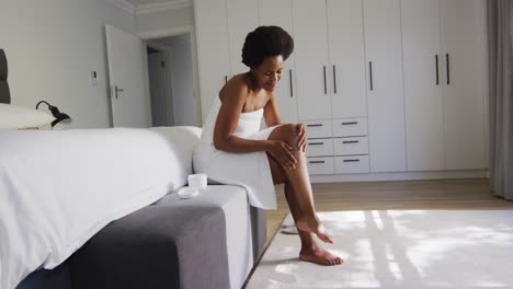 Happy-african-american-woman-apply-cream-on-legs-in-bedroom