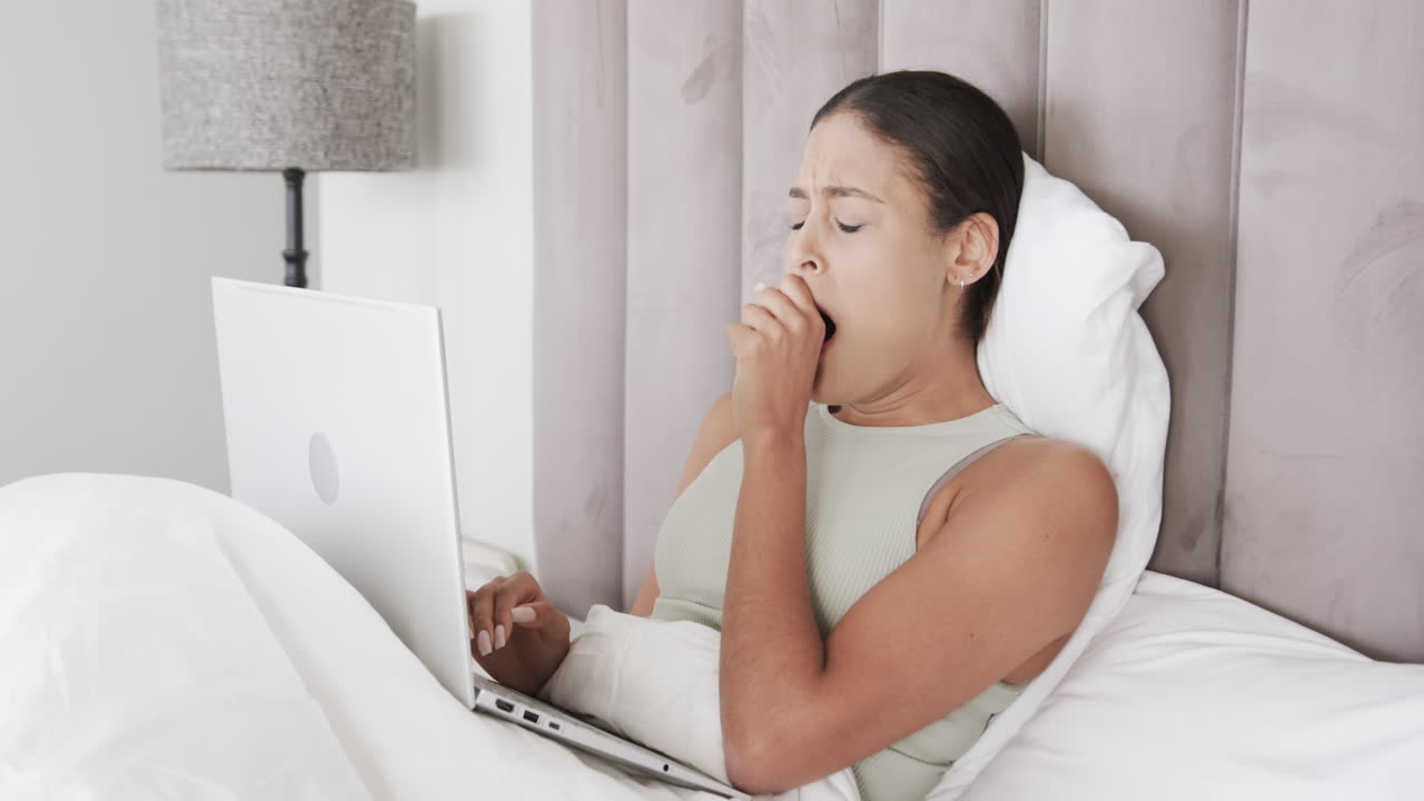 Biracial woman sitting on bed using laptop and yawning, slow motion