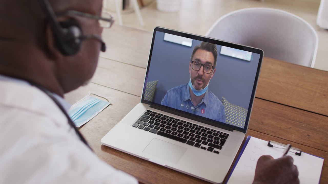 Ein afroamerikanischer Arzt mit Telefon-Headset macht sich Notizen während eines Videoanrufs am Laptop.