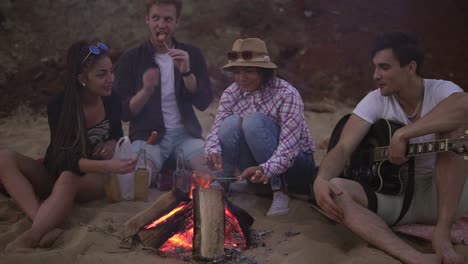 Group-of-young-and-cheerful-people-sitting-by-the-fire-on-the-beach-in-the-evening,-grilling-sausages-and-playing-guitar