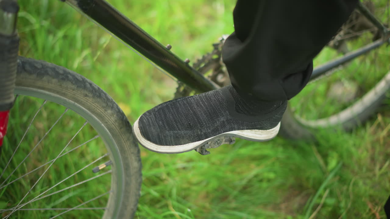 primer plano de un individuo con zapatillas grises tocando y girando el pedal de una bicicleta estacionada en un campo de hierba mientras toca su tobillo, el pie permanece en el pedal mientras la bicicleta se ajusta