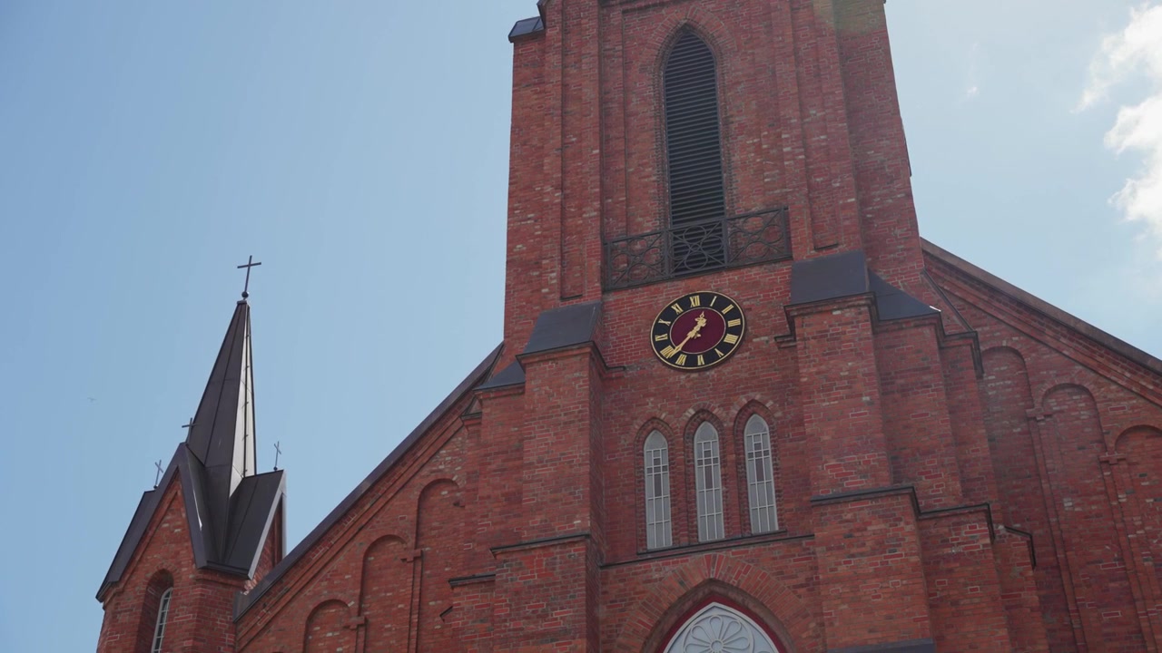 Ampia inquadratura statica della facciata neogotica in mattoni rossi della Cattedrale di Kaišiadorys in Lituania sotto un cielo azzurro brillante, dettagli architettonici e torre dell'orologio illuminati dalla luce solare naturale, nessuna persona visibile.