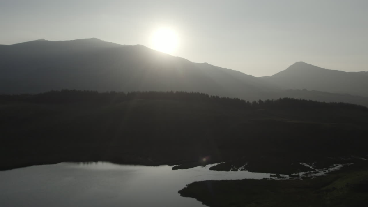 Reverse drone flyover of Llyn y Dywarchen lake at sunrise with Snowdon mountain in the distance. Scenic aerial footage captures morning light over North Wales countryside and tranquil rural landscape
