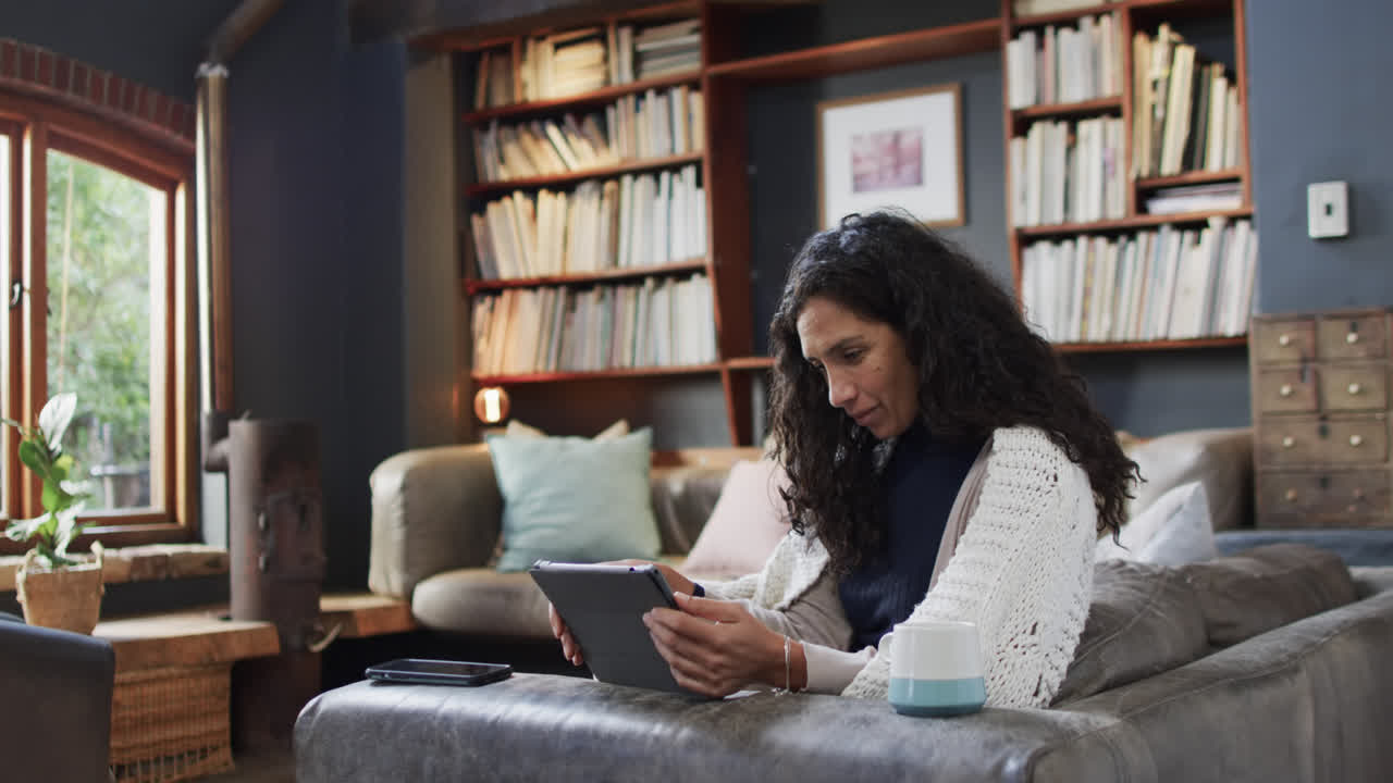 mujer biracial sentada en el sofá y usando la tableta en casa, cámara lenta con espacio de copia