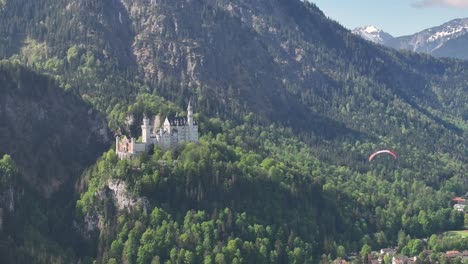 Statische-Aufnahme-Eines-Gleitschirms,-Der-über-Den-Rahmen-Fliegt,-Mit-Einer-Weißen-Burg-Im-Grünen-Wald-Und-Einem-Berggipfel-Im-Hintergrund