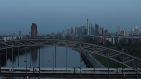 Frankfurt-am-Main-Skyline-at-Blue-Hour-early-morning-over-Main-River-with-passanger-Train-passing-bridge,-Aerial-high-angle-slow-forward