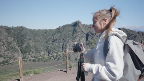 Vista-Lateral-De-Una-Linda-Joven-Turista-Con-Mochila-Sosteniendo-Una-Cámara-Tomando-Fotos-En-La-Naturaleza-Del-Campo-En-Un-Agradable-Día-Soleado