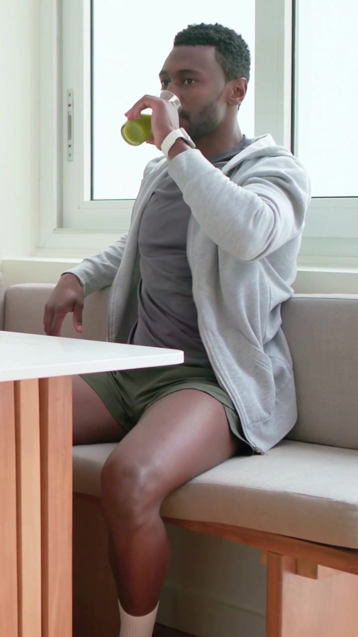 Vertical video: Placing glass, man reaching, sipping green juice and talking in home nook, hoodie