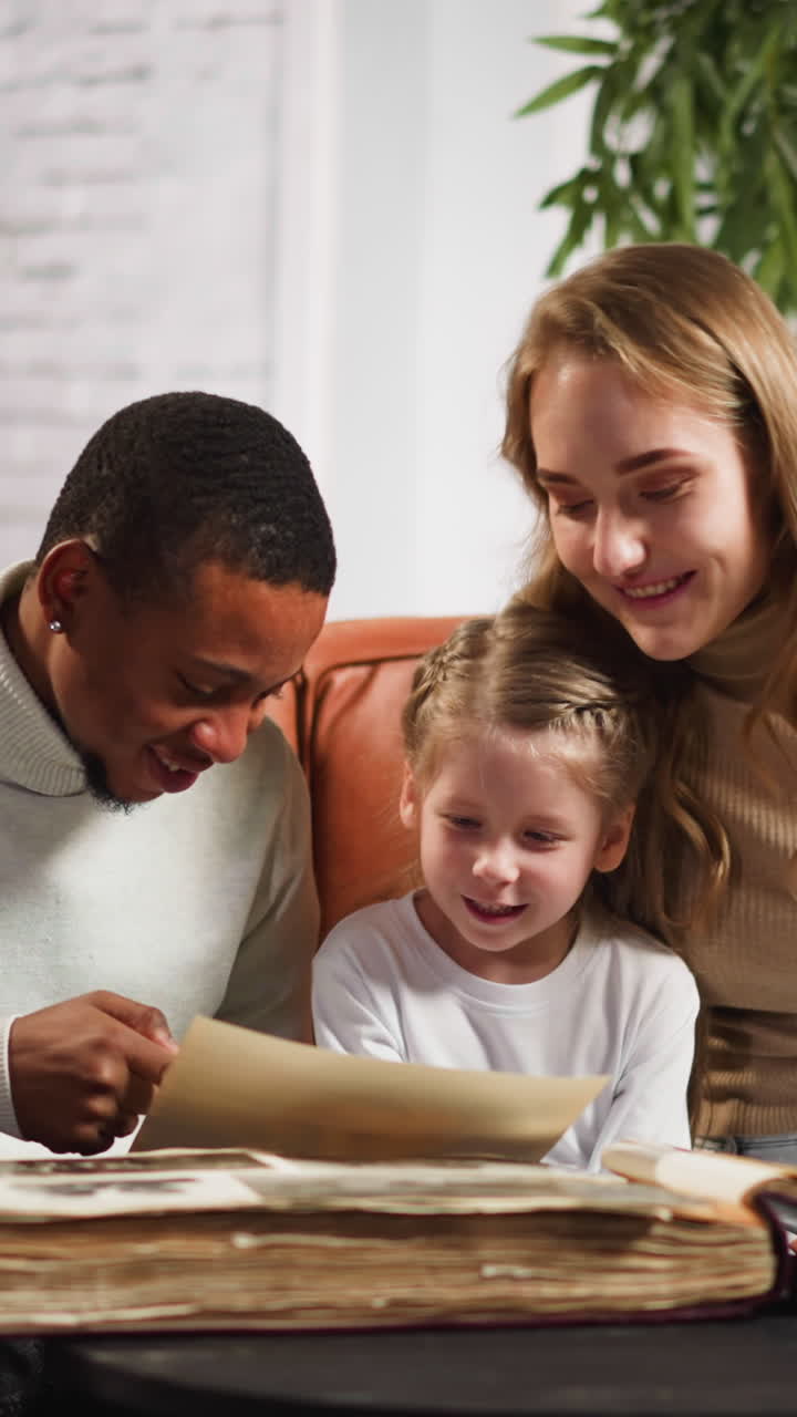 Black father shows photo of grandparents to happy mixed race daughter in old album sitting with blonde wife on sofa in living room slow motion