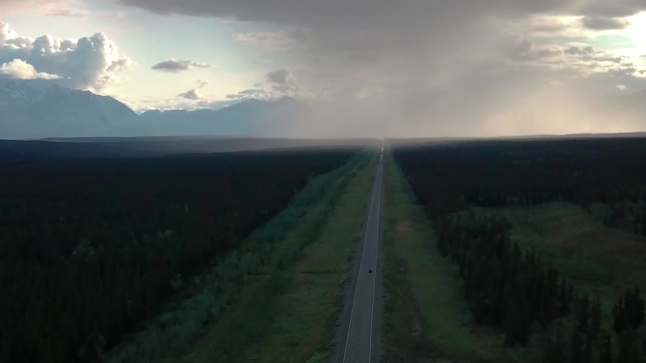 Sensational panoramic summertime scene above straight narrow countryside Alaskan highway in Ibex valley by evergreen trees in forest towards mountain range and rain clouds, overhead circle aerial