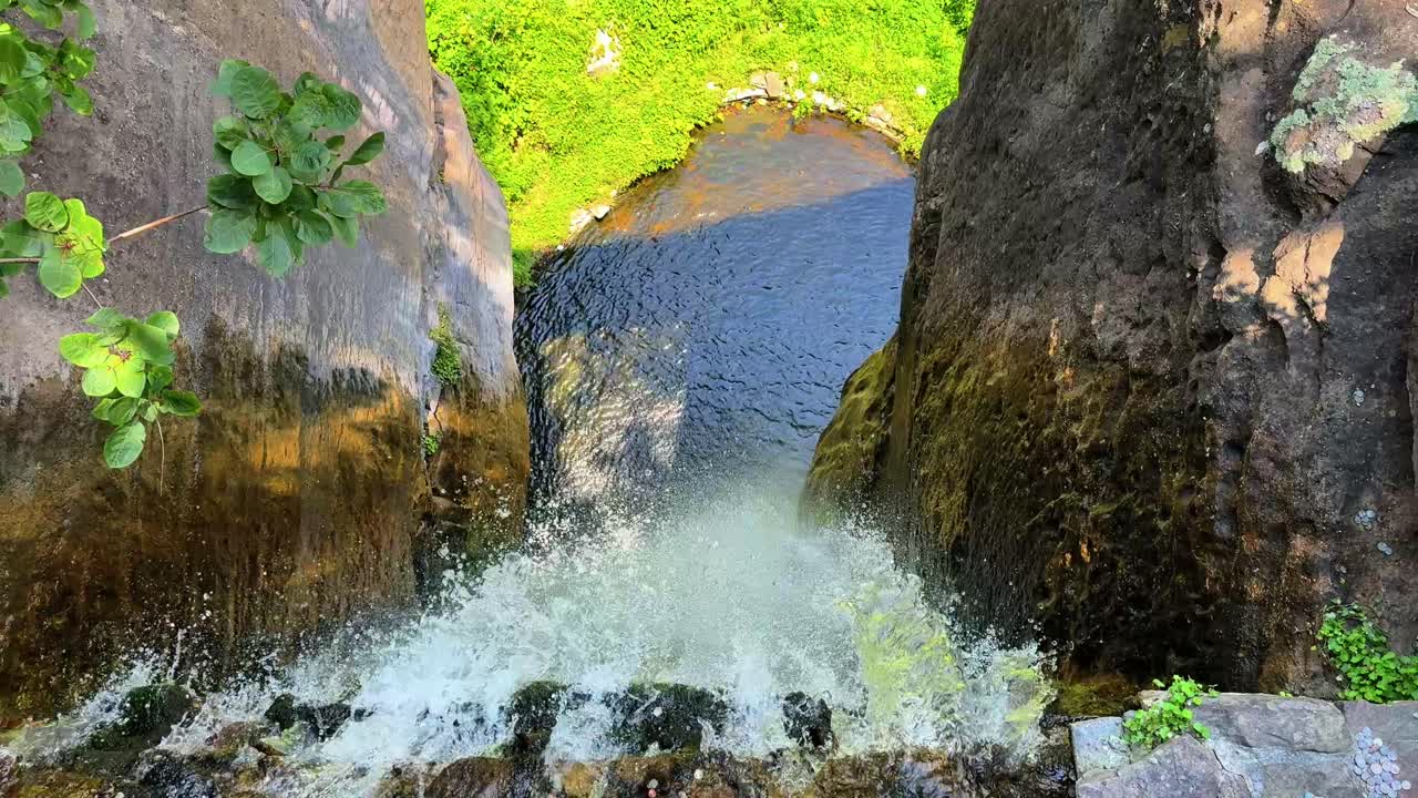 Experience an exciting first-person view of water rushing over a rocky cliff into a serene pool below. Vibrant greens surround, with bright, natural sunlight illuminating the scene