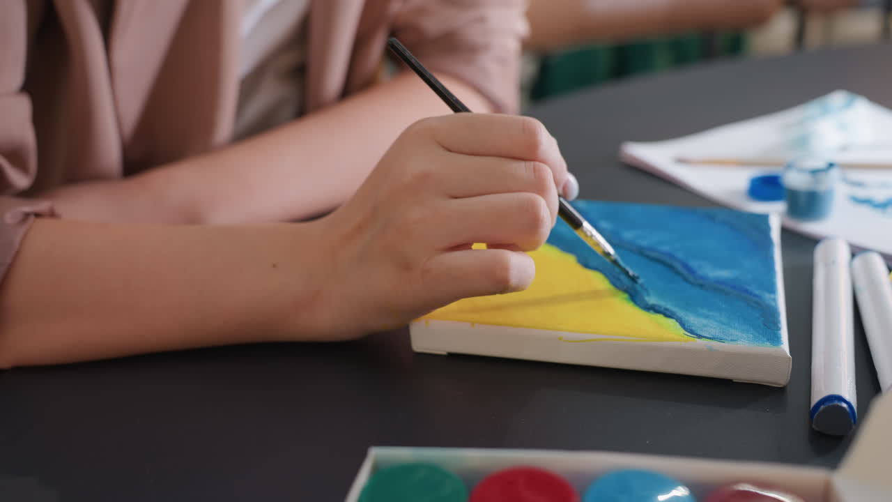 Primer plano de un pincel que añade un contorno ondulado sobre un lienzo pequeño, la mano del artista estabiliza el trazo, azul vibrante superpuesto sobre una base amarilla, paleta y rotuladores cerca, creación deliberada de textura para dar profundidad