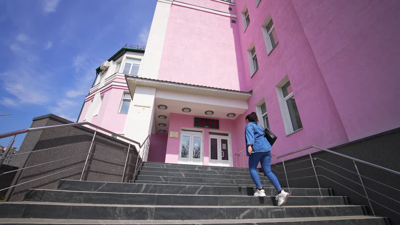 Coming up by the stairs with silver handrails to the doors of the big building. Following the girl in jeans wear going up by the staircase. Low angle view.