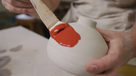 close up view of female potter wearing apron using glaze brush to paint on pot at pottery studio