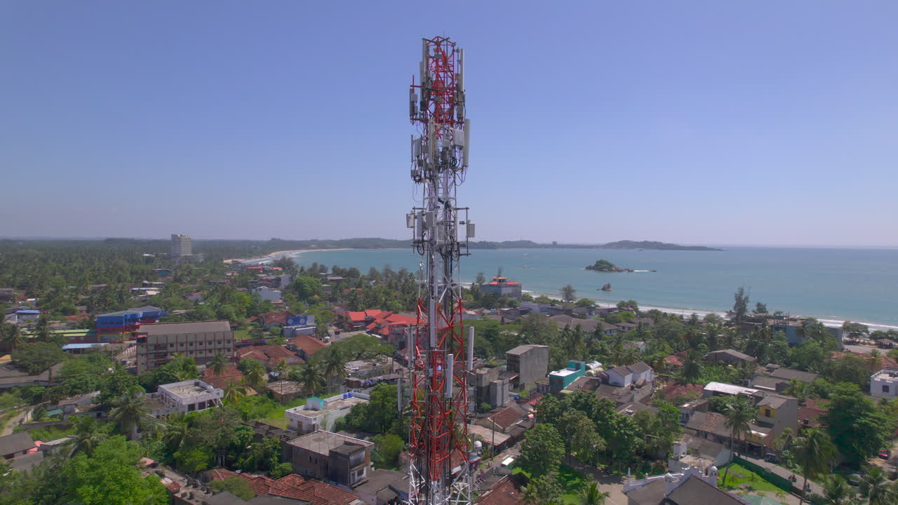 Torre di telecomunicazioni si erge sopra una città costiera tropicale con tetti di palme e una baia oceanica blu. Infrastruttura di rete mobile in un insediamento marittimo sotto la luce del giorno