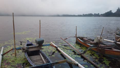 Wooden-Boats-Docked-Around-Lotus-Lilies,-Beratan-Lake-Bali-Indonesia-Cloudy-Sky-Fog-and-Mist-around-Scenic-Landscape