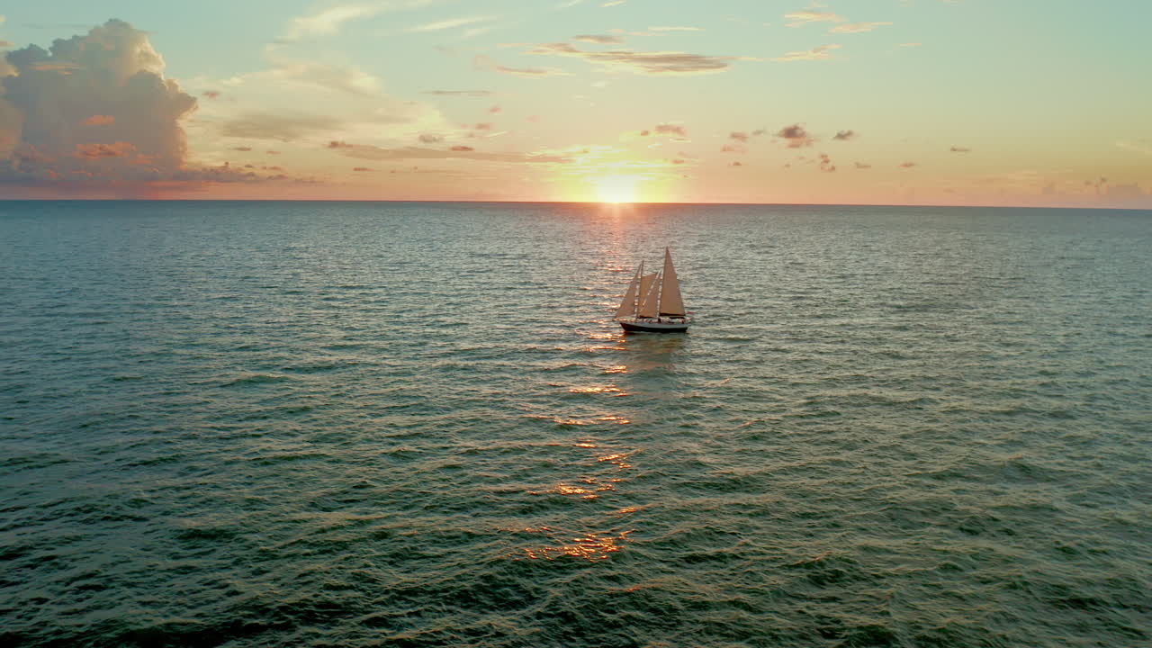 A sailboat glides across the ocean under a glowing sunset, its silhouette framed by golden light reflecting on the water and clouds along the distant horizon