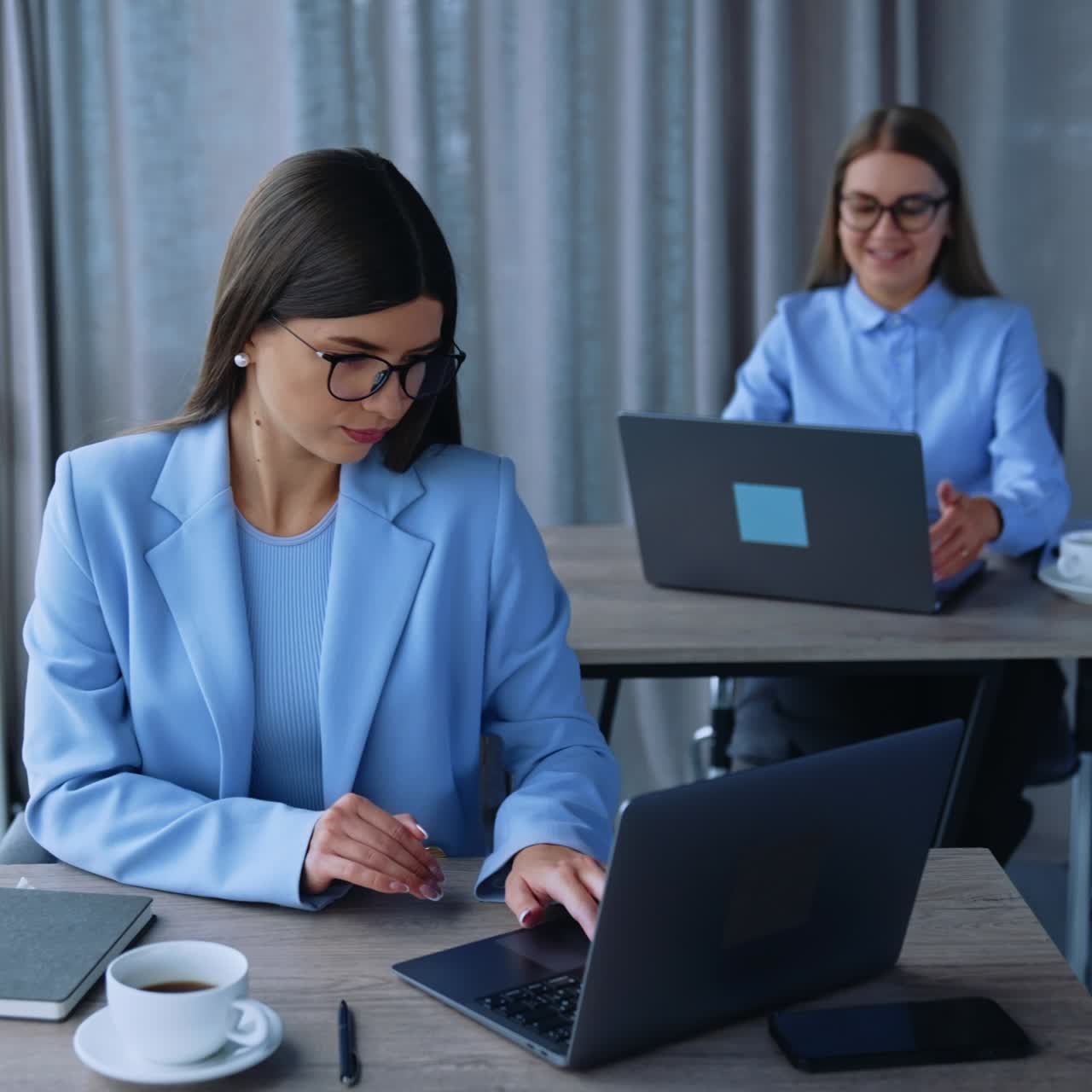 Two long-haired women wearing glasses sitting at desks in the office. Dark-haired lady opens her laptop and then paper notebook and takes a pen. Female colleague working at the computer at backdrop