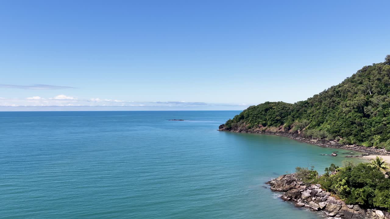 Drone pans above lush rainforest headland and turquoise sea under bright midday sunlight, clear sky