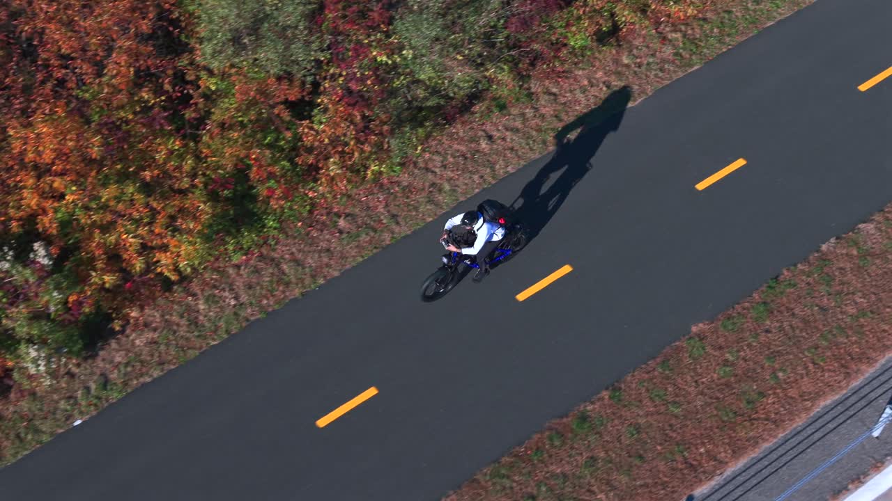 An aerial view of a man riding a bicycle on a paved bicycle path on a sunny day in autumn. The drone camera tilted downward, truck left and pan right following the rider
