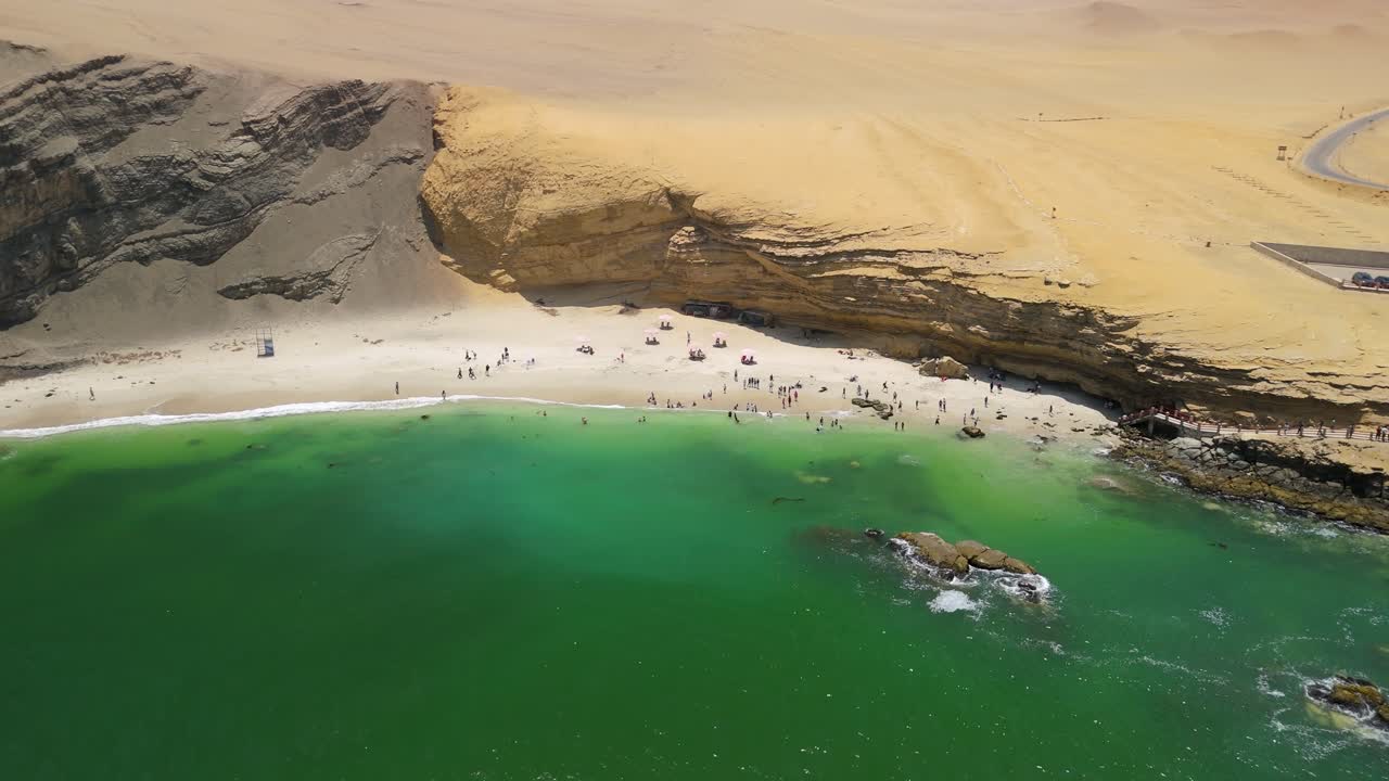 Aerial shot pulling back from the crowded public beach of Playa La Mina revealing tourists swimming in turquoise water at the base of desert cliffs