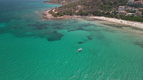 Hermosa-Costa-Con-Playa-Y-Hoteles-En-El-Fondo-De-La-Montaña-Con-Agua-Cristalina,-Gente-Nadando-Y-De-Pie-En-La-Playa-En-Cerdeña,-Italia---Tiro-De-Gran-Angular-De-Drones