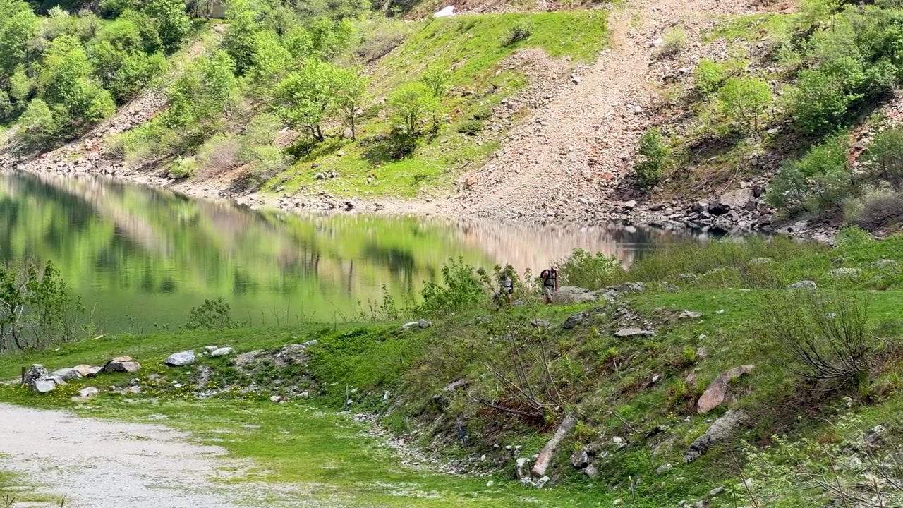 एक शांत झील के किनारे एक हरे भरे पहाड़ी रास्ते पर ट्रेकिंग करते हुए दो पैदल यात्रियों का वाइड शॉट।