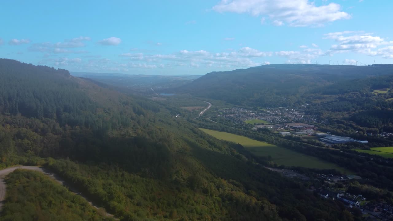 Aerial of Deep Welsh Valley with Winding Gravel Track on Mountain Side with Resolven Town with Open Landscape in Background. Factory Buildings in Rural Community with Blue Sky