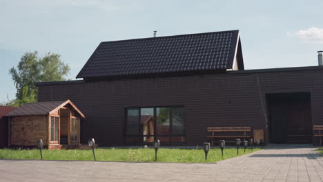 brown wooden house in sunny fenced compound with small wooden cabin, paved walkway leading to bench seating, green lawn swaying in breeze, simple landscape design under clear sky