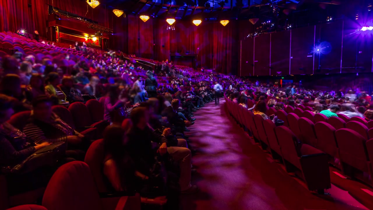 Penonton berkumpul di auditorium dan menyaksikan pertunjukan di teater dalam rekaman selang waktu. Aula besar dengan kursi berlengan merah. Penonton memenuhi tempat duduk hingga lampu dimatikan. Dilihat dari sisi kiri.