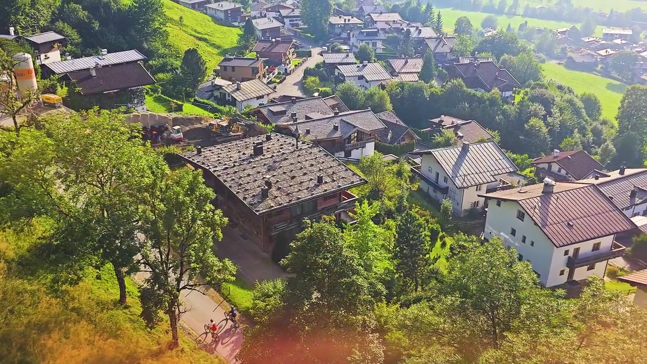 Prachtige bewegende gondelopname boven een alpine resortdorp, met groene dalweiden, verspreide huizen, beboste hellingen en wazige bergen in warm gouden zonlicht