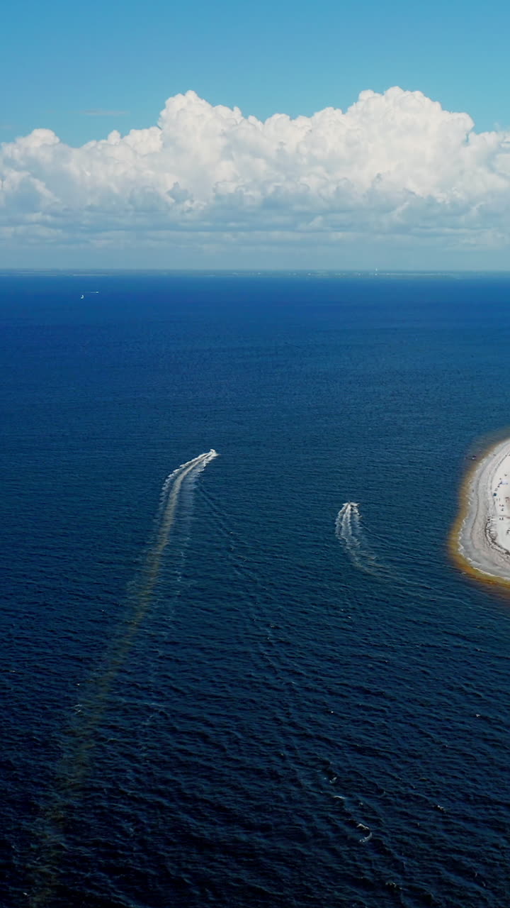 Two boats speed away from the coastline, carving white trails over deep blue waters as the sandy shoreline curves along the edge under a bright sky