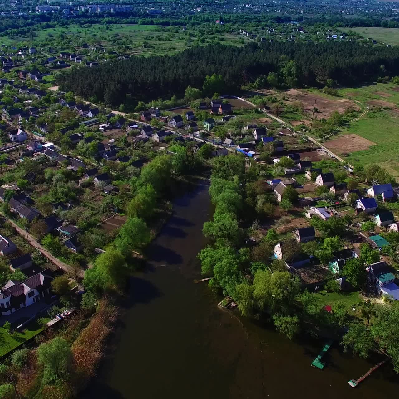 Panorama of the beautiful green village on the river bank. Flight over the shining water on sunny day. Big city at backdrop
