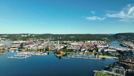 Kristiansand in Norway showcases a vibrant marina filled with boats, surrounded by lush green hills and buildings nearby, all under a clear, sunny blue sky
