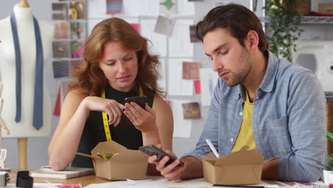 diseñadores de moda masculinos y femeninos teniendo almuerzo de trabajo mirando teléfonos móviles en el estudio