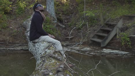 black man in a forest sitting on a tree trunk bridge over a creek