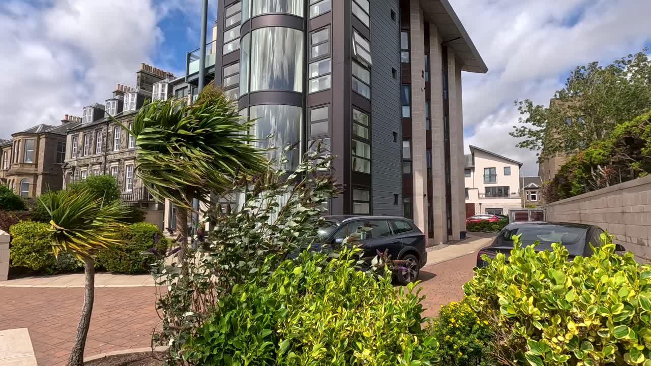 Un'auto di colore scuro percorre una strada residenziale passando accanto a un moderno condominio, circondata da una vegetazione lussureggiante, sotto la luminosa luce del giorno con un movimento di telecamera stabile.