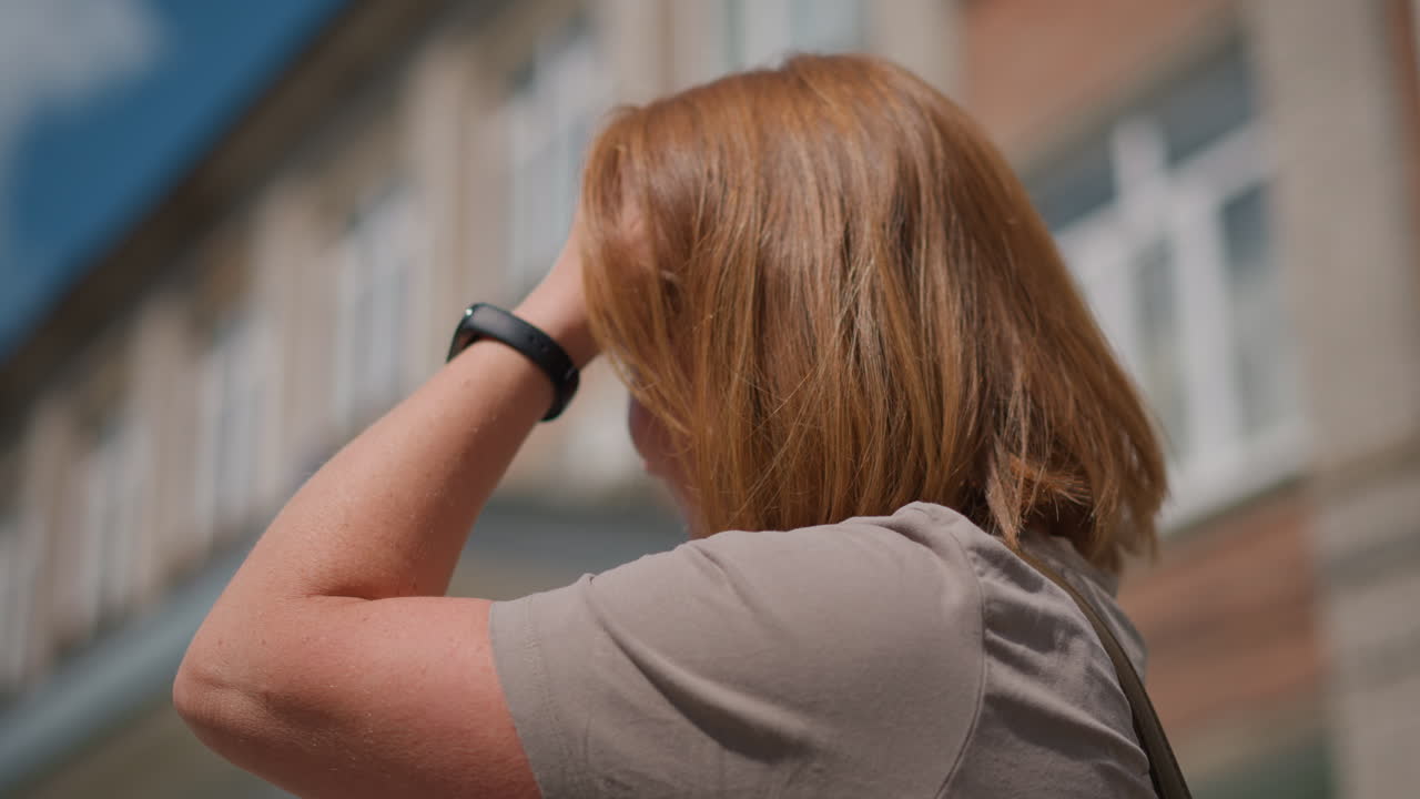 Side view of uneasy pupil adjusting hair with hand wearing smart watch, standing near residential building in daylight, expression shows discomfort and uncertainty under warm sunlight