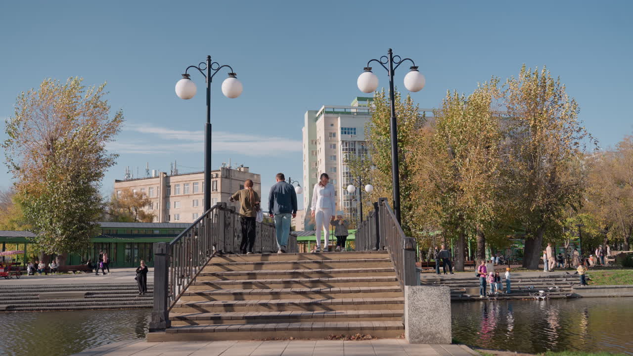 Coppia sposata che scende le scale di un ponte in un parco, skyline della città, alberi autunnali, lampioni, riflessi sull'acqua, luce solare calda, conversazione spontanea, passo tranquillo, atmosfera familiare romantica.