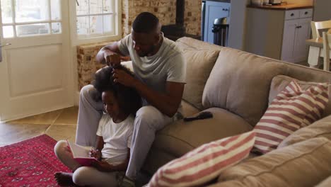 Afroamerikanischer-Vater,-Der-Seiner-Tochter-Die-Haare-Bindet,-Während-Er-Zu-Hause-Auf-Der-Couch-Sitzt