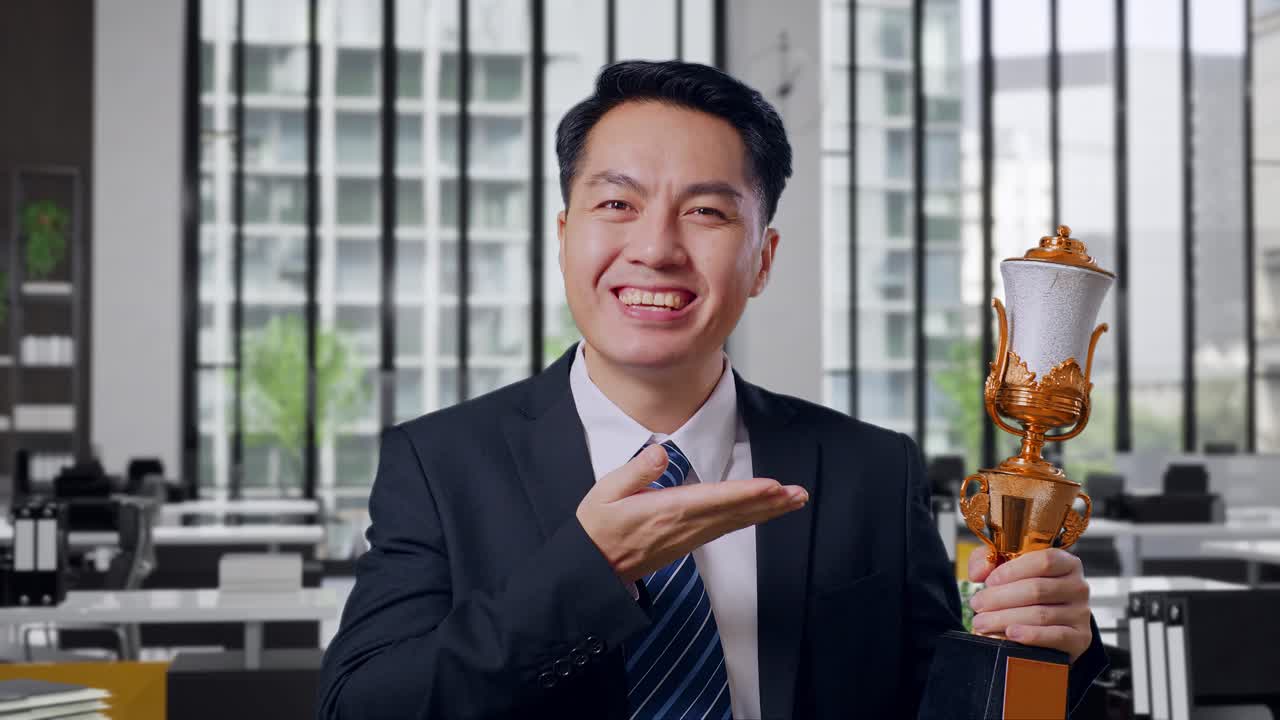 Close Up Of Asian Business Man In A Suit And Tie With A Gold Medal Pointing To A Gold Trophy In His Hand And Smiling Being Proud Winning As The First Winner In Office