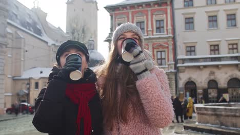 Two-smiling-women-tourists-traveling-together,-drinking-hot-tea,-coffee-from-cups-on-city-street